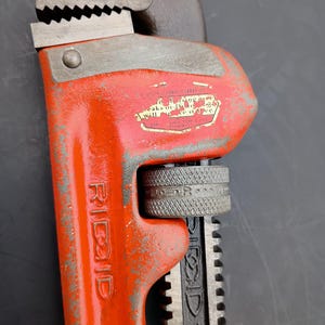 May include: Close-up of a Ridgid pipe wrench. The wrench is painted bright orange with the brand name in white. The adjustable jaw is gray and has sharp teeth. The handle is black and has the size "10" printed on it.