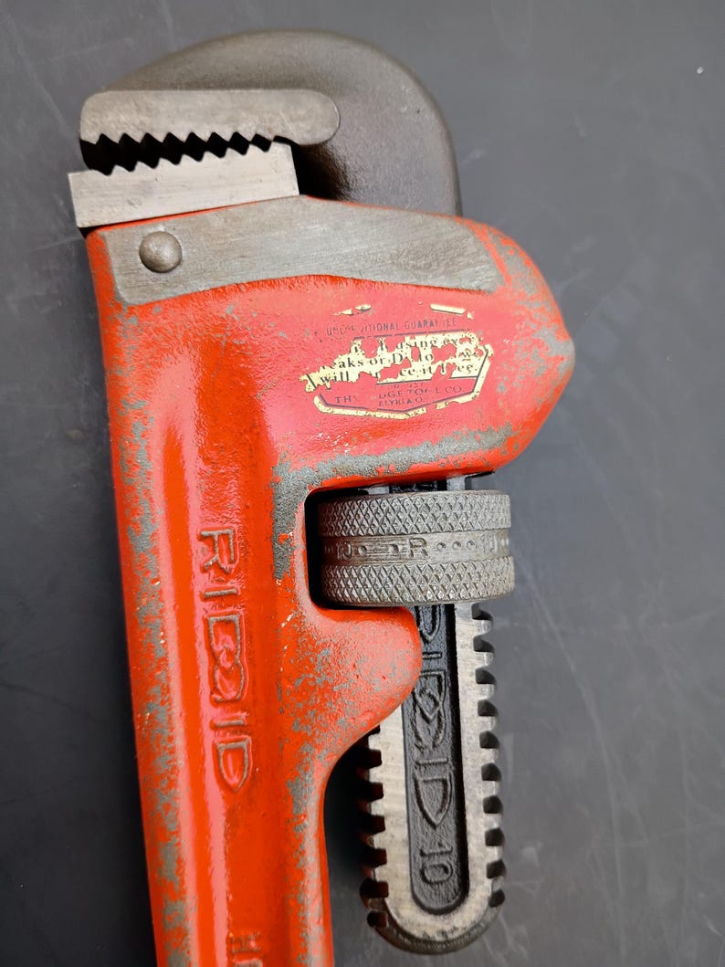 May include: Close-up of a Ridgid pipe wrench. The wrench is painted bright orange with the brand name in white. The adjustable jaw is gray and has sharp teeth. The handle is black and has the size "10" printed on it.