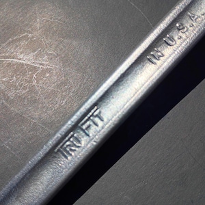 Op de afbeelding: Close-up van een zilveren moersleutel met de tekst "IRU FIT" en "IN U.S.A." in reliëf. De moersleutel staat schuin over een donkergrijze ondergrond, met schaduwen.