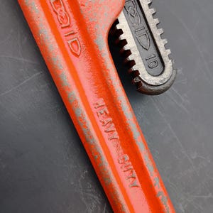 May include: Close-up of a heavy-duty RIDGID pipe wrench. The wrench is painted bright orange with the text "HEAVY DUTY" and "RIDGID" visible. The adjustable jaw is dark gray and shows the number "10".