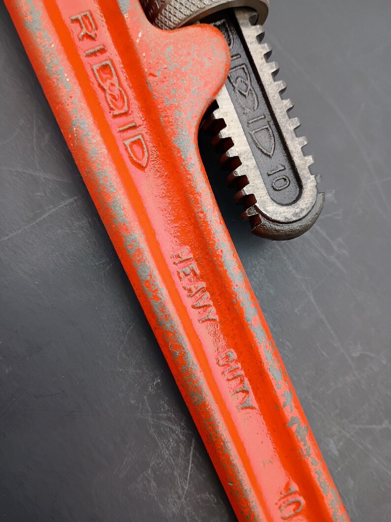 May include: Close-up of a heavy-duty RIDGID pipe wrench. The wrench is painted bright orange with the text "HEAVY DUTY" and "RIDGID" visible. The adjustable jaw is dark gray and shows the number "10".