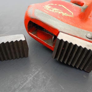 May include: Close-up of a red pipe wrench with a silver jaw and a silver threaded fitting. The wrench has a textured handle and a toothed jaw for gripping pipes. The tools are on a gray surface.
