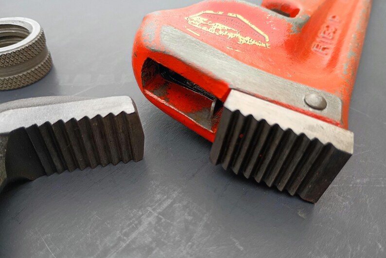 May include: Close-up of a red pipe wrench with a silver jaw and a silver threaded fitting. The wrench has a textured handle and a toothed jaw for gripping pipes. The tools are on a gray surface.
