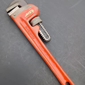 May include: An orange Ridgid pipe wrench, model 10, with a black adjustable jaw. The wrench is made of metal and shows signs of wear. The handle has a hole at the end for hanging. The wrench is designed for plumbing and mechanical work.