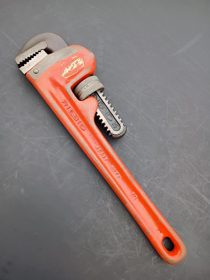 May include: An orange Ridgid pipe wrench, model 10, with a black adjustable jaw. The wrench is made of metal and shows signs of wear. The handle has a hole at the end for hanging. The wrench is designed for plumbing and mechanical work.