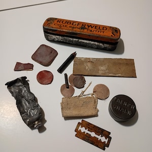 May include: An assortment of vintage items, including a metal tin labeled "RUBBERWELD CYCLE REPAIR OUTFIT", a crushed metal tube, a razor blade, and various small, circular objects. The items are arranged on a white surface.