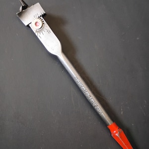 Puede incluir: Una broca de pala Irwin No. 21-USA A de color plateado con punta roja. La broca tiene una punta de tornillo y una hoja plana con marcas de medición. La herramienta está diseñada para carpintería y taladrado.