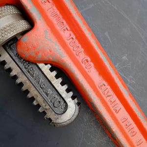 May include: Close-up of a vintage orange pipe wrench. The wrench has the text "THE RIDGE TOOL CO. ELYRIA OHIO U.S.A." and "WARREN, OHIO" printed on it. The wrench is a rusty orange color with some silver metal parts.