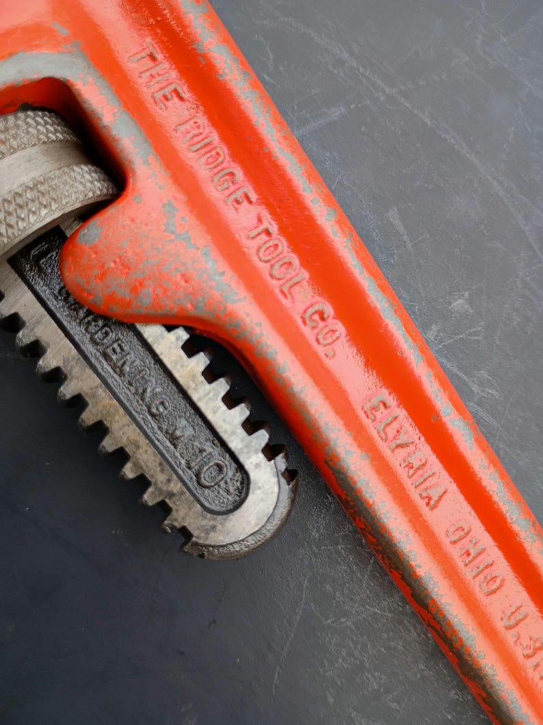 May include: Close-up of a vintage orange pipe wrench. The wrench has the text "THE RIDGE TOOL CO. ELYRIA OHIO U.S.A." and "WARREN, OHIO" printed on it. The wrench is a rusty orange color with some silver metal parts.