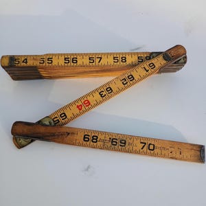 May include: A vintage wooden folding ruler, partially open, displaying inch measurements. The ruler is made of light-colored wood with dark brown markings and brass hardware. The numbers are printed in black and red.