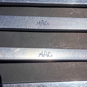 May include: Close-up of several silver metal bars with the embossed letters "MAC". The bars are rectangular and have a slightly textured surface. The image is taken in natural light, with a dark background.