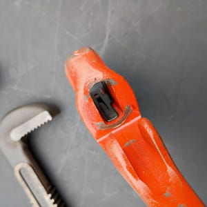May include: An overhead shot of a red and gray pipe wrench with a silver fitting on a gray surface. The wrench is open, showing its adjustable jaws. The fitting is a small, circular piece with a threaded interior.
