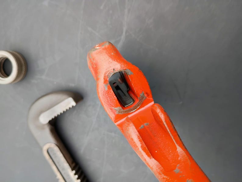 May include: An overhead shot of a red and gray pipe wrench with a silver fitting on a gray surface. The wrench is open, showing its adjustable jaws. The fitting is a small, circular piece with a threaded interior.