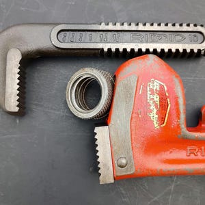 May include: A close-up of a RIDGID 10 pipe wrench in black and red, with a silver metal fitting. The wrench has a toothed jaw and the brand name is visible. The background is a dark gray surface.