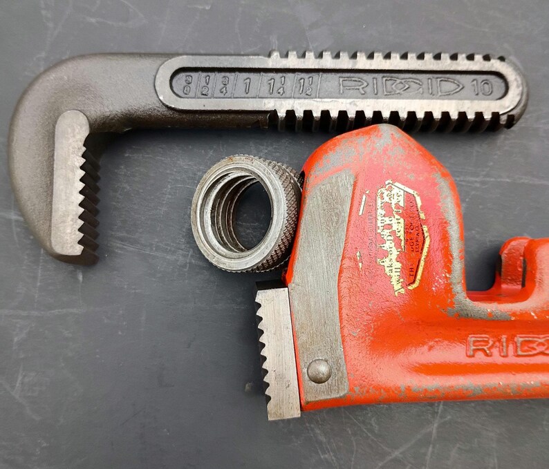 May include: A close-up of a RIDGID 10 pipe wrench in black and red, with a silver metal fitting. The wrench has a toothed jaw and the brand name is visible. The background is a dark gray surface.