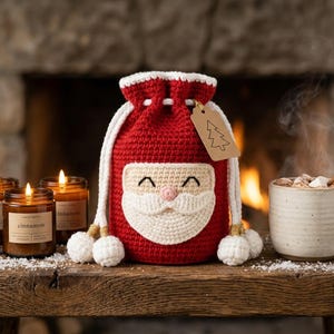 May include: A red and white crocheted drawstring bag featuring a Santa Claus face design on the front. The bag has a white drawstring with pom-pom ends and a brown paper gift tag with a line drawing of a Christmas tree attached to the side.