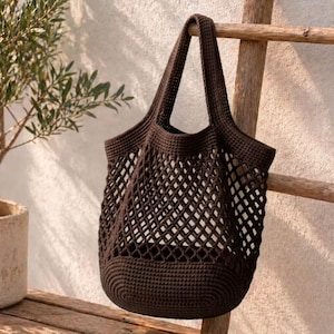 Puede incluir: Un bolso tote de ganchillo marrón oscuro con un diseño de red. El bolso tiene dos asas y está colgado de una escalera de madera. El bolso está hecho de hilo grueso y tiene una base redondeada. Ideal para compras.