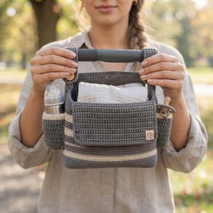 May include: A dark gray and cream striped crochet caddy with a sturdy gray handle, a front flap pocket, and two side compartments. The caddy contains a white cloth, a silver baby bottle, and a pacifier. A small rectangular label is attached to the front.