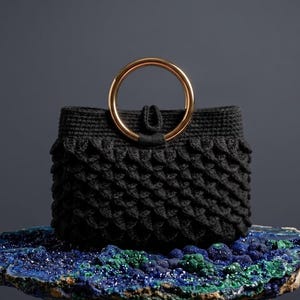 May include: A black crocheted handbag featuring a textured scale-like pattern and a single circular gold-toned metal handle. The bag has a structured rectangular body with a small central loop closure at the top, showcasing a detailed yarn construction.