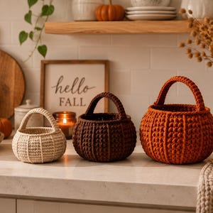 Può includere: Tre cesti rotondi all'uncinetto a forma di zucca, disposti in fila. I cesti sono color crema, marrone scuro e arancione bruciato, ognuno caratterizzato da un esterno testurizzato e a coste e da un singolo manico arcuato sul bordo.