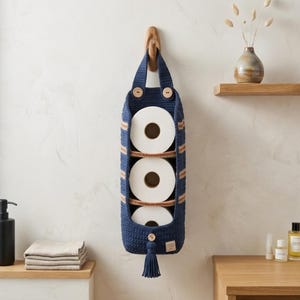 May include: A navy blue crocheted hanging storage organizer featuring three vertical compartments holding white toilet paper rolls. The organizer has a top handle with two wooden buttons, a bottom tassel, and a small rectangular label on the front.