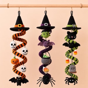 Puede incluir: Tres decoraciones colgantes en espiral de ganchillo para Halloween suspendidas de una varilla de madera. La izquierda presenta rayas naranjas y negras con calaveras, una calabaza y un murciélago. El centro tiene espirales moradas con un monstruo, murciélago, caldero y araña. La derecha tiene rayas verdes y negras con un gato, murciélago, dulces de maíz, una lápida y una araña.