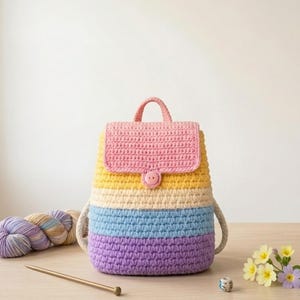 May include: A crochet backpack featuring horizontal stripes in pink, yellow, cream, light blue, and purple. The bag has a top handle, a flap closure secured with a single pink button, and thick white rope straps attached to the sides.