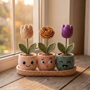 Könnte beinhalten: Drei gehäkelte Blumen in einzelnen Töpfen mit gestickten Gesichtern, angeordnet auf einem passenden ovalen Tablett. Die Blumen umfassen eine beige Tulpe, eine gelbe Rose und eine violette Tulpe, jeweils mit grünen Blättern in Garn-Töpfen.