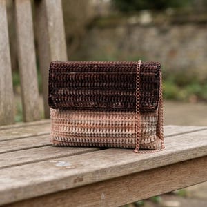 May include: A rectangular crochet handbag featuring a dark brown upper flap and a light pink and beige gradient lower section. The bag is constructed with a textured, ribbed stitch pattern and includes a thin, metallic rose gold chain shoulder strap.