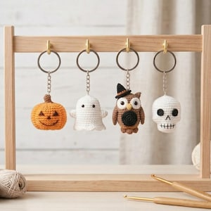 May include: Four crochet keychains hanging from a wooden stand. The charms include an orange pumpkin with a face, a white ghost, a brown owl wearing a black witch hat, and a white skull with black eye sockets and a stitched mouth design.
