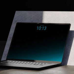 May include: A black laptop with a dark screen displaying the time 10:13 in light blue digits. The device features a thin bezel, a standard keyboard layout, and a sleek, dark finish, positioned at an angle against a light grey rectangular panel.