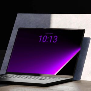 Könnte beinhalten: Ein schwarzer Laptop mit einem dunklen Bildschirm, der den violetten Text 10:13 anzeigt. Der Bildschirm weist einen leuchtend violetten Lichteffekt am rechten Rand auf und das Gerät ist vor einer hellgrauen, schrägen Fläche platziert.