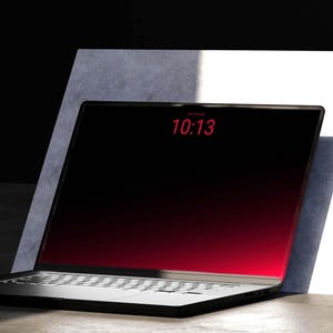 Könnte beinhalten: Ein schwarzer Laptop mit einem rot verlaufenden Bildschirm, der die Uhrzeit 10:13 anzeigt. Der Laptop ist geöffnet und angewinkelt und lehnt an einer weißen und grauen rechteckigen Struktur. Die Tastatur ist silber.