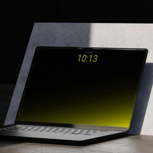 Könnte beinhalten: Ein schwarzer Laptop mit einem hellen Bildschirm, der die Uhrzeit 10:13 anzeigt. Der Laptop ist geöffnet und abgewinkelt, an eine weiß-graue strukturierte Oberfläche gelehnt. Der Bildschirm hat einen gelben Farbverlauf.