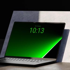 Könnte beinhalten: Ein schwarzer Laptop mit einem leuchtend grünen Bildschirm, der die Uhrzeit 10:13 und den Text 94% Charged anzeigt. Der Laptop verfügt über ein schmales Rahmendesign und eine Standardtastatur und steht vor einer hellen, schrägen Fläche.