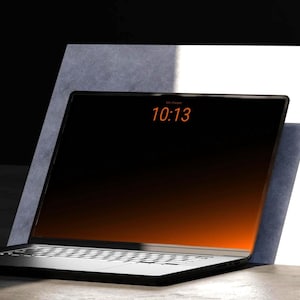 Könnte beinhalten: Ein schwarzer Laptop mit weißer Tastatur und einem Bildschirm, der die Uhrzeit 10:13 in Orange anzeigt. Der Laptop ist an einer weißen und grauen Oberfläche angelehnt, mit dunklem Hintergrund.