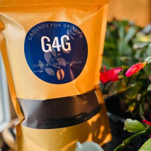 May include: A tan paper bag of coffee grounds with a dark brown band across the middle. A blue circular label reads "Grounds for Growth" and "G4G" with the text "Support the Spectrum" and a coffee bean graphic. The bag is surrounded by green plants and red flowers.