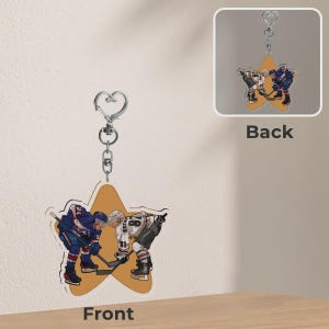 Op de afbeelding: Een stervormige sleutelhanger met een hockeythema. De voorkant toont twee hockeyspelers in actie, met een gele sterachtergrond. De achterkant van de sleutelhanger toont ook een hockeytafereel. De sleutelhanger heeft een zilverkleurige sluiting.