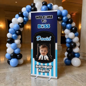Peut inclure: Une bannière verticale avec le texte Welcome To Boss et Birthday Party ainsi qu'une photo d'un bébé en costume. La bannière est encadrée par une arche de ballons bleus, blancs et noirs. Le fond de la bannière est bleu foncé avec des rayures bleu clair.
