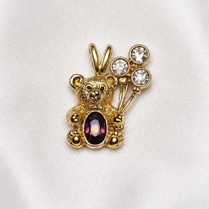 May include: A gold-toned pendant shaped like a teddy bear holding three round clear crystals on thin stems. The bear features a textured finish and a large, oval-shaped deep purple gemstone set in its center, with a bail at the top for a chain.