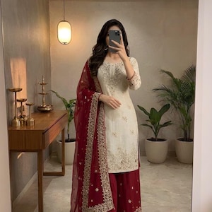 May include: Cream-colored embroidered kurta and palazzo set with a maroon dupatta. The outfit features intricate gold embroidery and is paired with matching maroon palazzo pants. The dupatta has a delicate, sheer appearance.