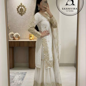 Op de afbeelding: Een witte geborduurde salwar kameez set met gouden versieringen. De outfit bevat een tuniek met lange mouwen, een broek met wijde pijpen en een bijpassende sjaal. Het Saanvira Store logo is zichtbaar.