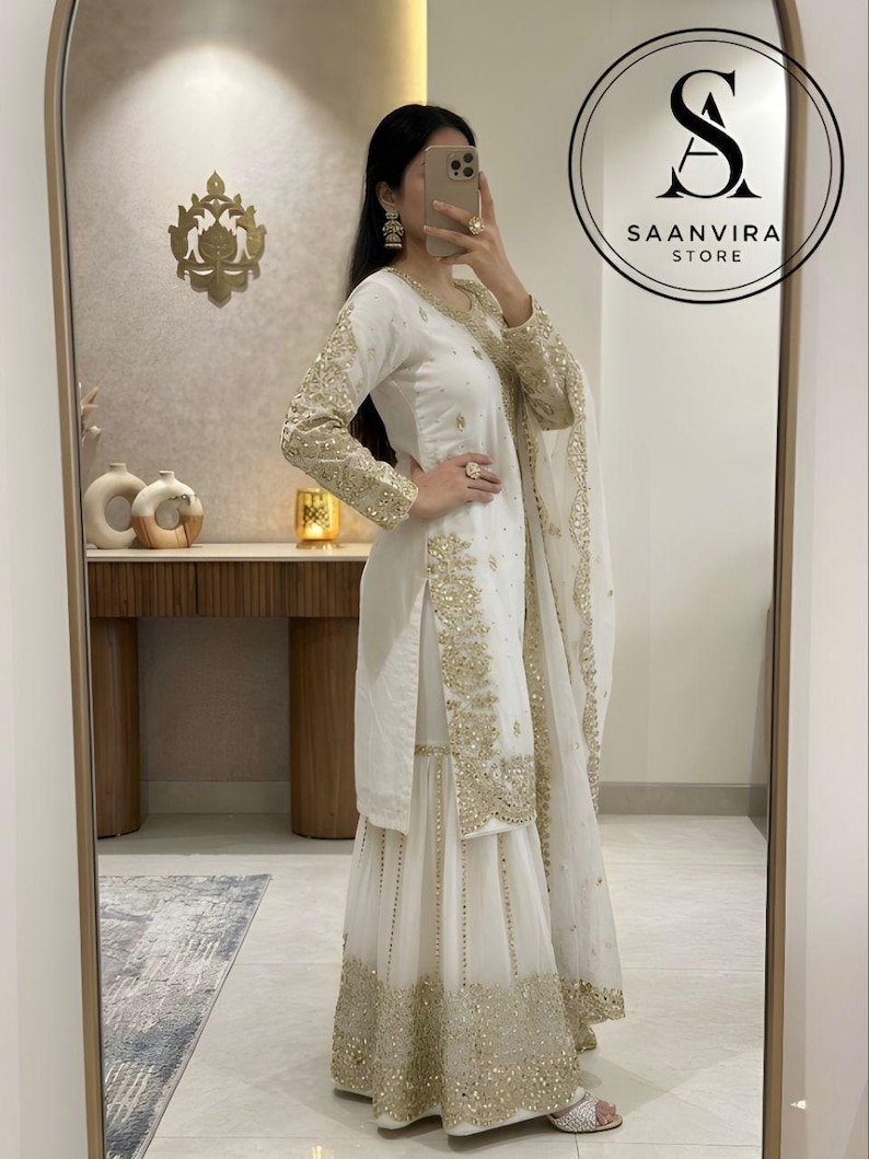 Op de afbeelding: Een witte geborduurde salwar kameez set met gouden versieringen. De outfit bevat een tuniek met lange mouwen, een broek met wijde pijpen en een bijpassende sjaal. Het Saanvira Store logo is zichtbaar.