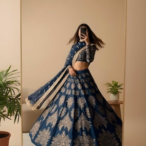 May include: A navy blue lehenga with intricate gold embroidery. The ensemble includes a long-sleeved, cropped top and a flowing skirt. A matching dupatta with gold trim completes the look. The image is reflected in a large mirror.