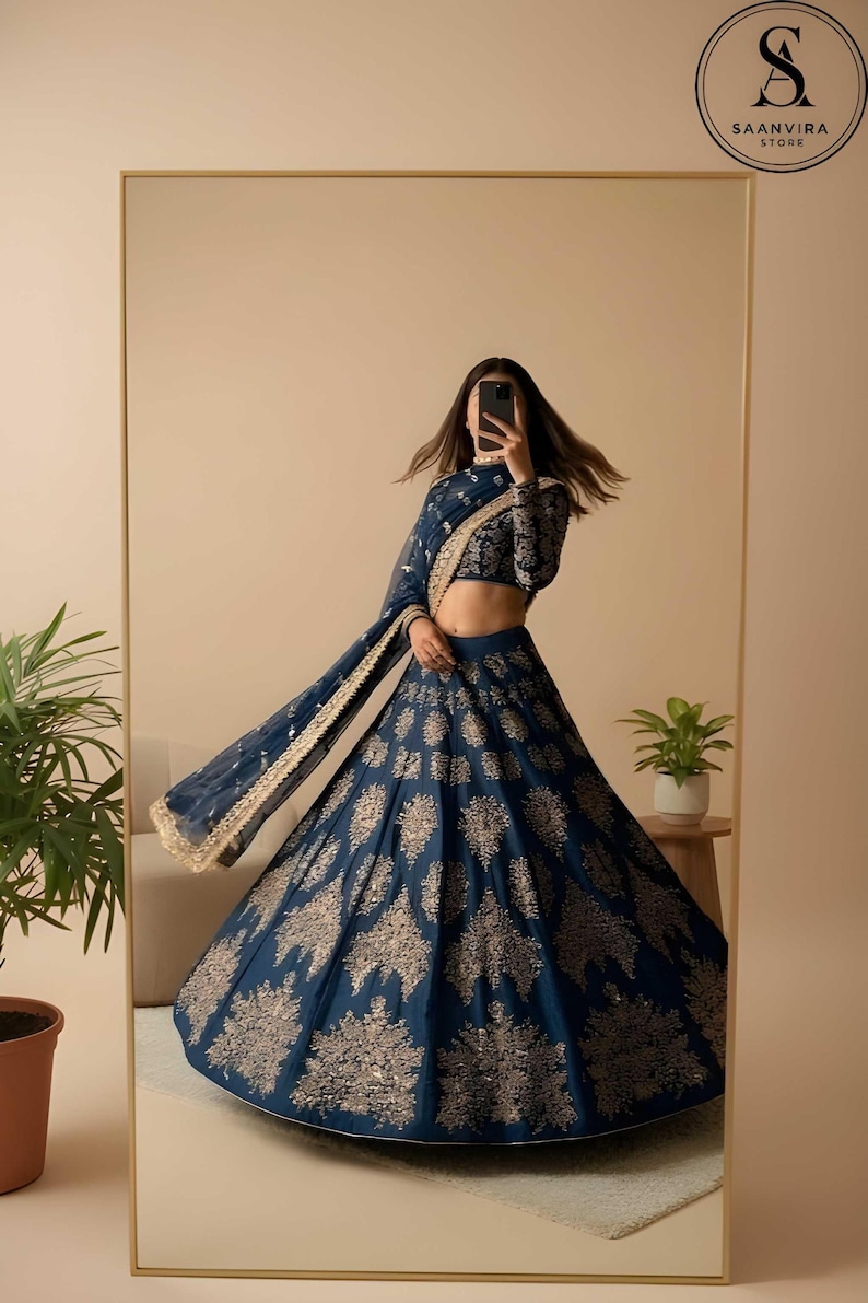 May include: A navy blue lehenga with intricate gold embroidery. The ensemble includes a long-sleeved, cropped top and a flowing skirt. A matching dupatta with gold trim completes the look. The image is reflected in a large mirror.