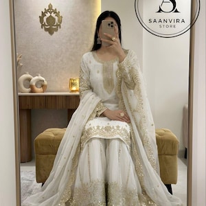 Op de afbeelding: Witte outfit met gouden details, bestaande uit een tuniek met lange mouwen, een broek met wijde pijpen en een bijpassende sjaal. De outfit heeft gedetailleerde gouden borduursels en pailletten. Het Saanvira Store logo is zichtbaar.