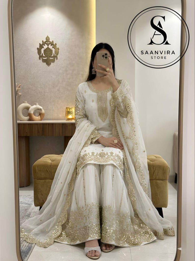 Op de afbeelding: Witte outfit met gouden details, bestaande uit een tuniek met lange mouwen, een broek met wijde pijpen en een bijpassende sjaal. De outfit heeft gedetailleerde gouden borduursels en pailletten. Het Saanvira Store logo is zichtbaar.