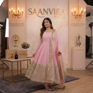 May include: A woman wearing a light pink Anarkali suit with gold sequin detailing. The suit includes a long-sleeved top, pants, and a matching dupatta. The background features the text "SAANVIRA STORE" and decorative lighting.