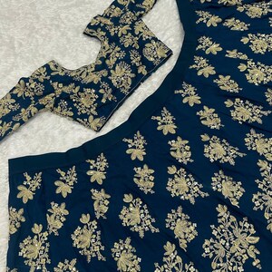 May include: A dark teal ensemble featuring intricate gold floral embroidery. The set includes a long-sleeved top, a skirt, and a matching scarf, all adorned with detailed golden patterns. The fabric has a subtle sheen.