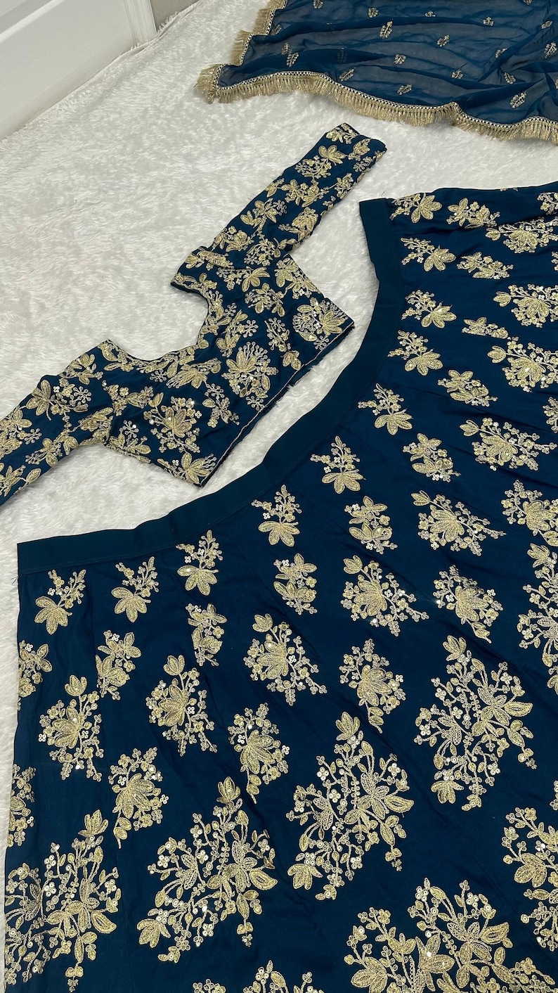 May include: A dark teal ensemble featuring intricate gold floral embroidery. The set includes a long-sleeved top, a skirt, and a matching scarf, all adorned with detailed golden patterns. The fabric has a subtle sheen.